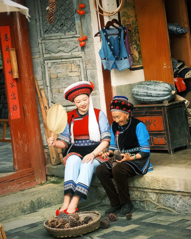 Two Bai women in traditional embroidered dress and headwear sitting on a doorstep with a basket of pine cones