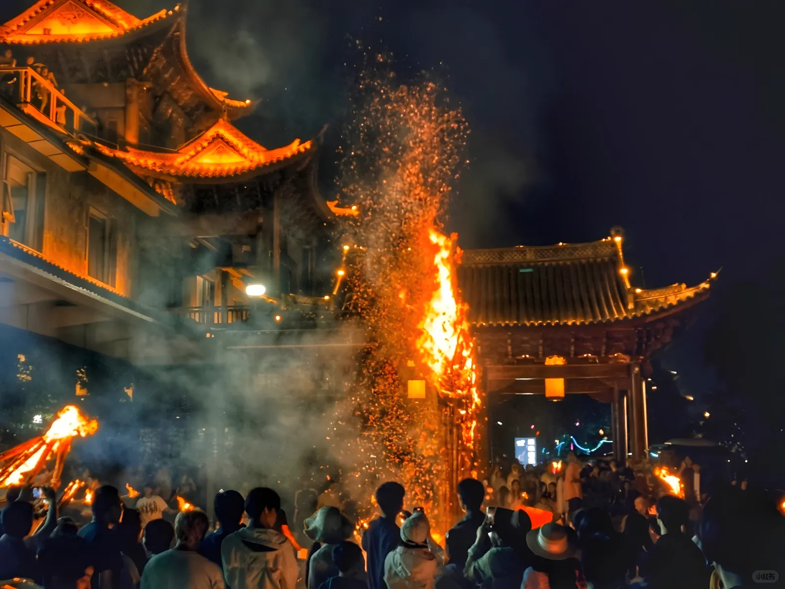 A towering torch showering sparks over a crowd beneath lantern-lit Bai gate towers on the night of the Torch Festival in Dali